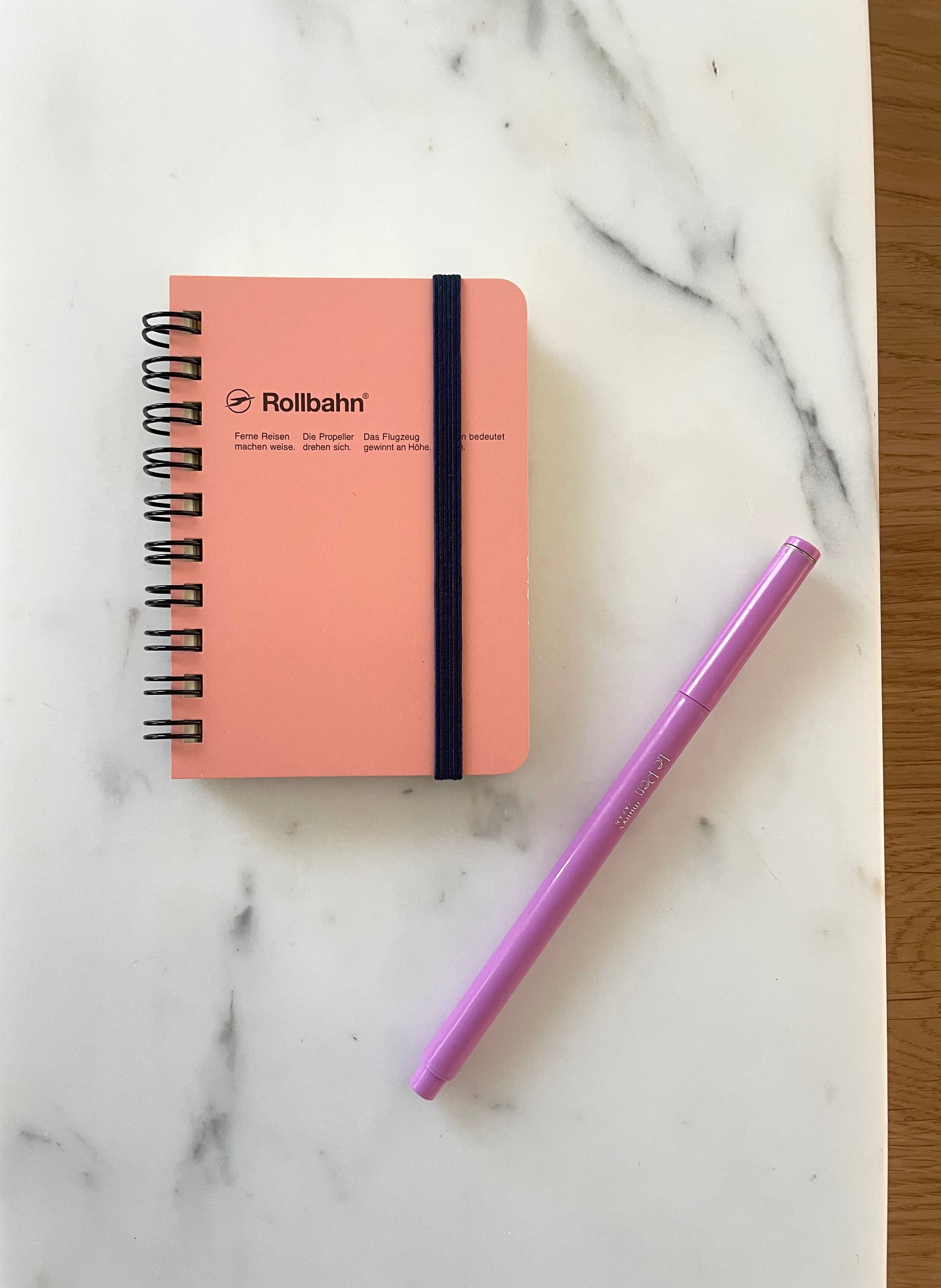 A blush pink Rollbahn spiral mini memo notebook with a grommeted elastic strap and a purple lilac Le pen lying in front of it, placed on a marble surface.